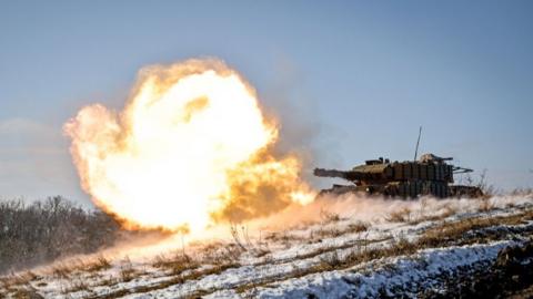 A tank participates in a live fire exercise. A large muzzle blast of fire can be seen shooting out of the tank's main gun.