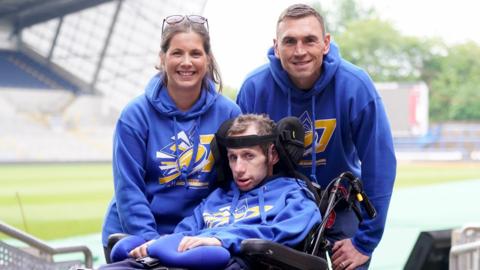 Kevin Sinfield, Rob Burrow and Lindsey Burrow look at the camera at a sport stadium. All three are wearing blue hooded tops. 
