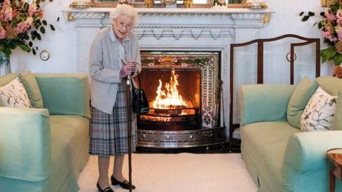 The late Queen Elizabeth stands in front of a fireplace at Balmoral. She has short white hair and wears glasses. She has a walking stick and wears a grey cardigan and a tartan pleated skirt