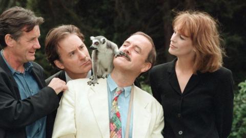 The cast of Fierce Creatures around John Cleese with a Lemur on his shoulder, placing a claw on the actor's mouth