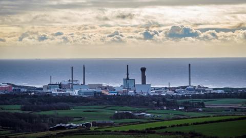 A general view of Sellafield taken from afar across a green field. The skyline of Sellafield is characterized by tall buildings and towers, with the sea in the background. 