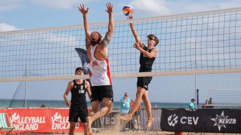 Three men playing beach volleyball. A man in black vest and shorts jumps to meet a multi-coloured ball behind a high net, while a man in white vest and black shorts jumps to defend in front of the net. A second man in black stands to the left. Spectators can be seen in the background in front of a blue sea.