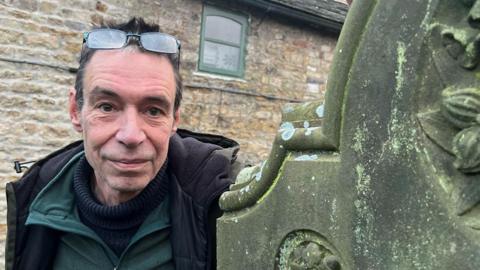 Close-up of Bob Sutton, wearing layers of warm clothing with his glasses on his head, leaning against a lichen-covered headstone