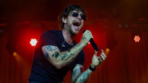 Tom Grennan singing on stage. He holds a microphone and his mouth is wide open. He wears dark glasses and a black T-shirt and has tattoos on his arms.