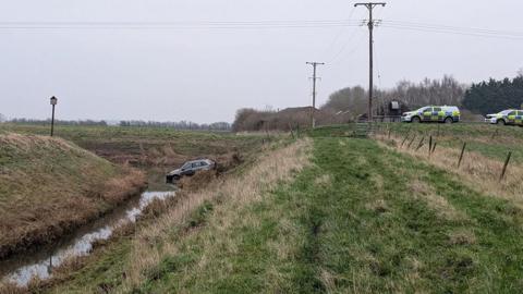 A view of a dark coloured car in a water filled ditch surrounded by fields with a number of police cars on the top of the bank