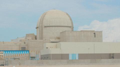 A large sand-coloured building in the UAE called the Barakah Nuclear Power Plant. In the centre is a large dome while rectangular shaped sections of the building lie to the left, right, and in front of the dome. The sky behind is blue with a few white clouds.