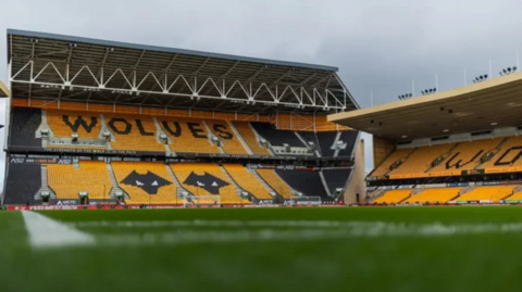 A view of two sides of Wolverhampton Wanderers football stadium and part of the green pitch. There are rows of orange seats in the stands with the word "Wolves" written out in black seats in the top tier. There are two black-coloured heads of wolves - the club's emblem - created with black seats in the bottom part of the stand.