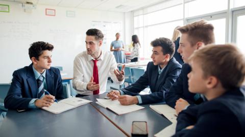 A young male teacher talks to a group of four boys in a classroom. The pupils are all sat around in navy blazers over light blue shirts and ties, while the teacher wears a white shirt with red tie.