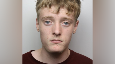 A police mugshot of a man with short blonde hair and a dark t-shirt.