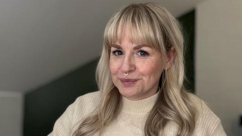 Katie is sitting at her desk. She is wearing a cream turtleneck jumper and has a nose ring. Her hair is long and a silvery, blonde colour, with a straight fringe. 