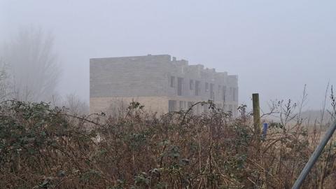 An unfinished two-storey block of flats in light and dark brick. There are no roof or windows and in the foreground are overgrown bushes 