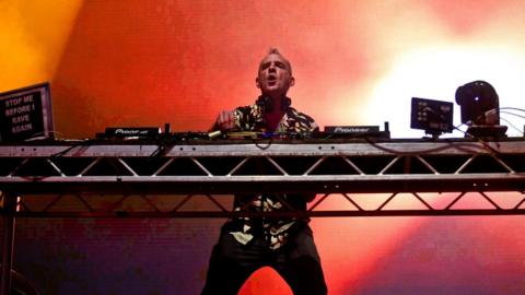 A man in a flowery short sleeved shirt stands behind a set of decks. Behind him is a pink and red screen with flashing lights.