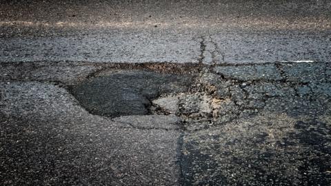 Close-up image of a pothole. It is roughly circular with a number of cracks, and part of it looks to have been filled with asphalt, which is darker in colour than the surrounding road surface.