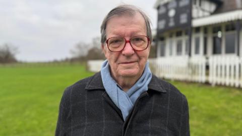 A man stood in a field with a building behind him which is black and white and surrounded by a fence and blurred. He has short grey hair, large red-rimmed glasses and a black blazer and light blue scarf on.