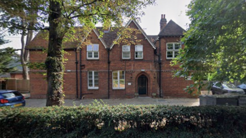 A red-brick building stands behind trees and bushes. The building has two apex roof sections and six large white-framed windows. It has a door arch for an entrance and a paved area out the front used as a car park.