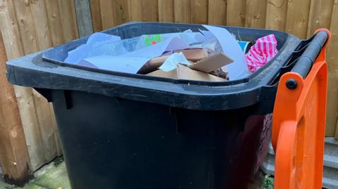 Dark grey wheelie bin with the orange lid opened, and paper and cardboard visible inside. The bin is in front of a wooden fence.