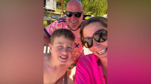 Three people pose for a selfie. A woman on the right is wearing sunglasses and a bright pink top. A boy is on the left. A man with a bald head and sunglasses is standing behind the pair. All of them are smiling into the camera. Green bushes, lawns and tables are behind them.