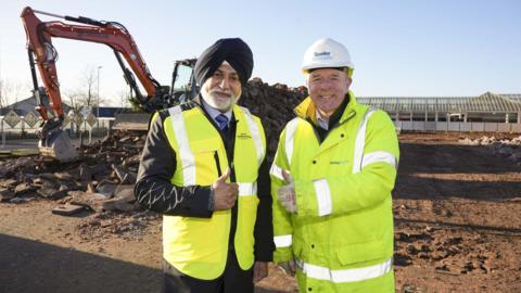 Two men are standing on a building site that has mud on the ground. They are wearing yellow hi-vis jackets and have their thumbs up. There is an orange digger in the background and a large market building.