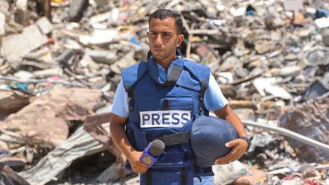 Anas al-Shariff, wearing a blue Press vest and carrying helmet and mic, stands in front of a pile of rubble, 2024