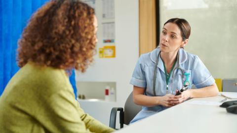 Female nurse in a blue uniform talking to a female patient wearing a green cardigan