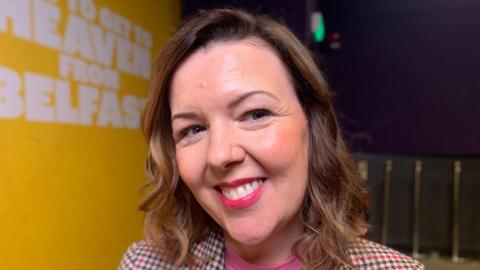A woman with brown hair and a checked jacket in front of a yellow background.