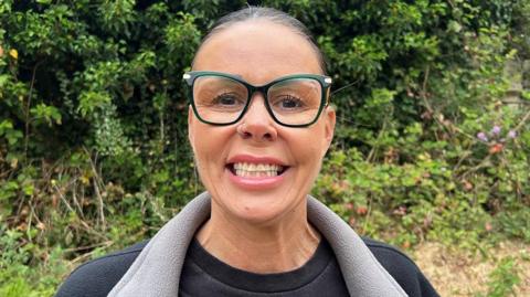 Head and shoulders image of Annette Shuttleworth. She is standing outside with greenery behind her. She is smiling, wearing large glasses and a black t-shirt and fleece. Her dark hair is tied back off her face