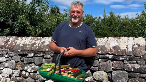 Adam Day, a man with grey hair and a beard, stands against a dry stone wall holding a basket containing vegetables from his allotment in Greystoke. He is wearing a dark t-shirt and smiling at the camera.