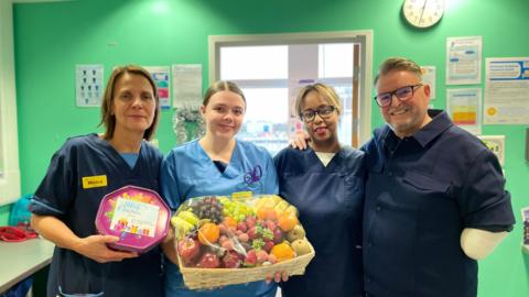 Matt stands on the right next to three nurses from ward 408 of the Queen Elizabeth Hospital. One nurse is holding a box of chocolates and another holds a fruit hamper.