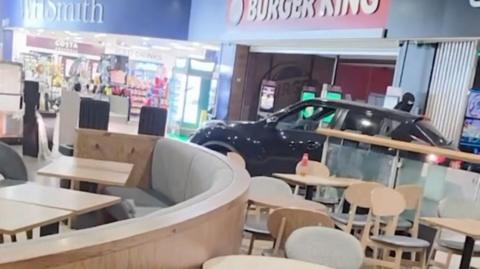 A black SUV has driven into a motorway service station. It is pictured on the other side of some cafe seating reversing through the location.