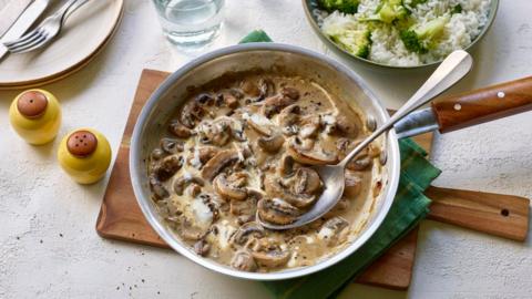 A silver saute pan filled with creamy mushrooms on a wooden board next to a bowl of rice and broccoli