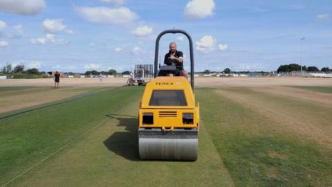 A man on a yellow cricket pitch roller with a parched outfield all around the square