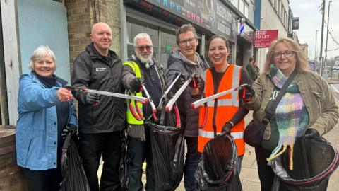 Luisa Scott among a group of five people with litter pickers and bin bags on Gateshead High Street. She has dark hair, tied back, and is wearing an orange hi-vis jacket. They are all looking directly into the camera and smiling.