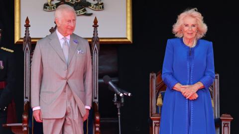 King Charles III and Queen Camilla pictured smiling at an event; the King wearing a grey suit and the Queen dressed in a bright blue coat.