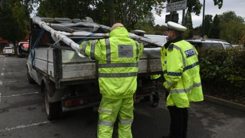 Two men in florescent jackets inspect a pick-up truck with a heavy load. One is a policeman and the other is from the Driver Standards Agency. The truck is white. 