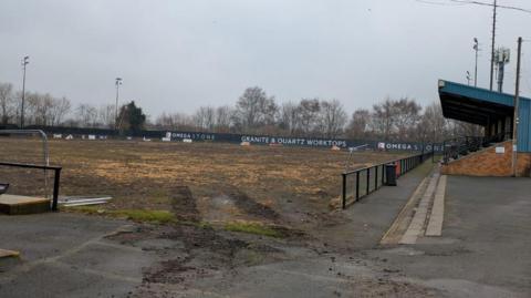 A pitch which is mostly mud, surrounded by advertising boards and lights. A spectator stand on one side of the pitch.