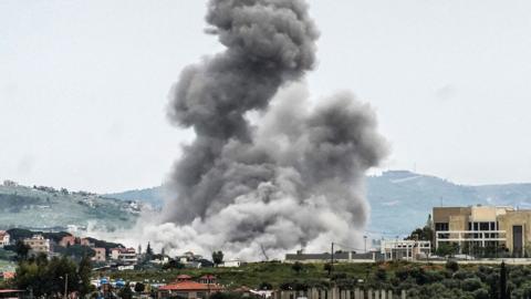 A large grey bundle of smoke rising from a village on a hillside. The smoke cloud fills most of the picture and is dramatic against the backdrop with village buildings in the foreground and hills in the background.