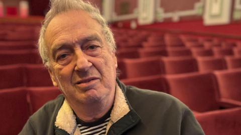 Film director Sir Stephen Frears sitting in the front row of an otherwise empty cinema. He is wearing a grey jumper over a black and white striped shirt.