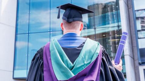 A graduate wearing a gown and mortarboard stands outdoors in front of a glass building, seen from behind, holding a rolled diploma. The gown is grey and he wears a green and purple hood on top. 