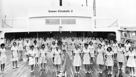 Dozens of women in their cleaning dresses pose with a vacuum cleaner on the deck of Queen Elizabeth 2 in 1975