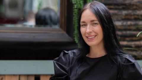 A woman wearing a black dress, with dark hair sitting on a wooden bench.