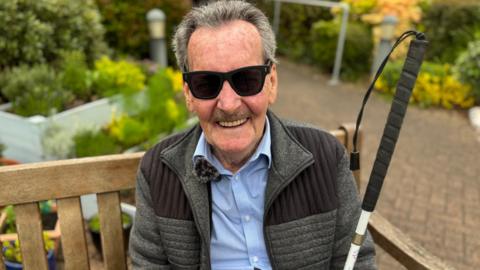 Jim Duffy sits on a wooden bench outside. He wears a grey jacket, blue shirt and sunglasses. He is smiling and has grey hair. There is a garden with paving stones and shrubs behind him.