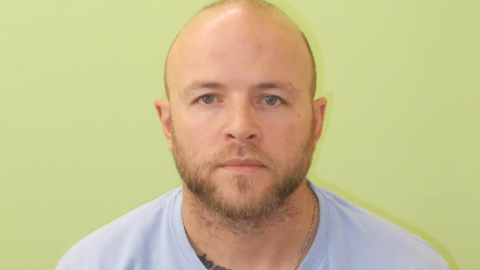 Kirk Bishop with short hair and beard wearing a pale blue T-shirt against a green background