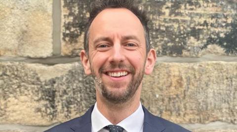 Ed Whiting smiles at the camera. He is standing against a stone wall and wearing a dark suit, white shirt and black tie with whit polka dots 