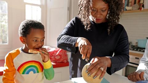 A mother in a kitchen cooking while her son next to her eats a piece of toast.