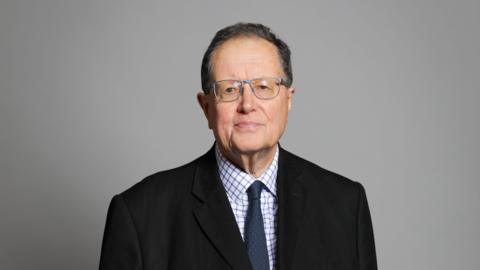Lord Boswell wearing a dark suit with a checked shirt and a tie. He is wearing light coloured glasses for his official House of Lords portrait.