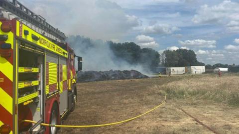 A fire engine is parked in a field with hoses unravelled as firefighters spray water on a straw fire as smoke fills the air with trees in the background