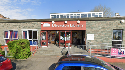 A single storey, grey-bricked building. The sign is red with white text. There are three steps leading up to the open doors, where book shelves can be seen beyond the threshold