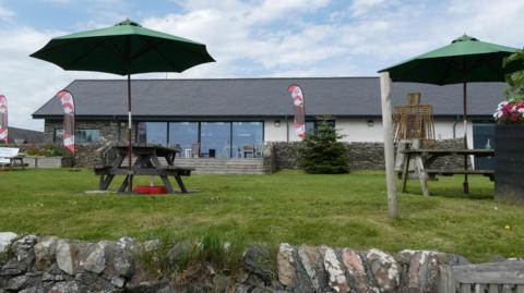 A tea room in Galloway with seats outside under umbrellas and a low-roofed building viewed across a grassy area.