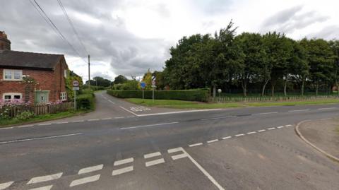 A Google street view image showing a crossroads junction. A cottage sits behind a fence on the left, behind pink flowers. A small wood is across the road on the right behind a hedge and wooden fencing. There are speed limit and other road signs by the road.
