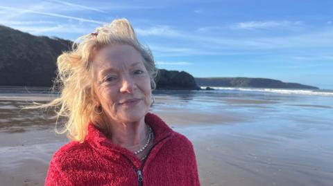 Kate Freeman is standing on the beach looking at the camera. Her long, blonde hair is being caught in the wind and she is wearing a red fluffy jumper. The sea can be seen in the background along with with the cliffs above. 